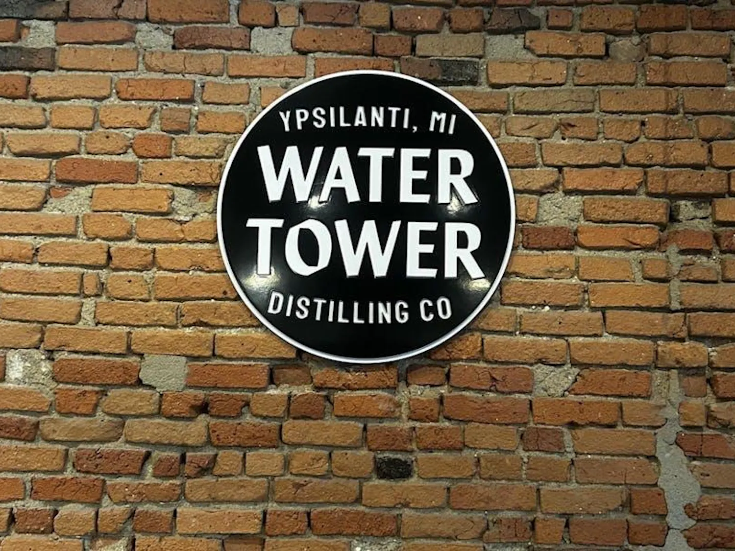 A red brick wall with a circular black sign hanging on it. There are four lines of white print on the sign. The first says "Ypsilanti, MI." The second says "Water," the third says "Tower," and the fourth says "Distilling Co." All of the letters on the sign are capitalized.