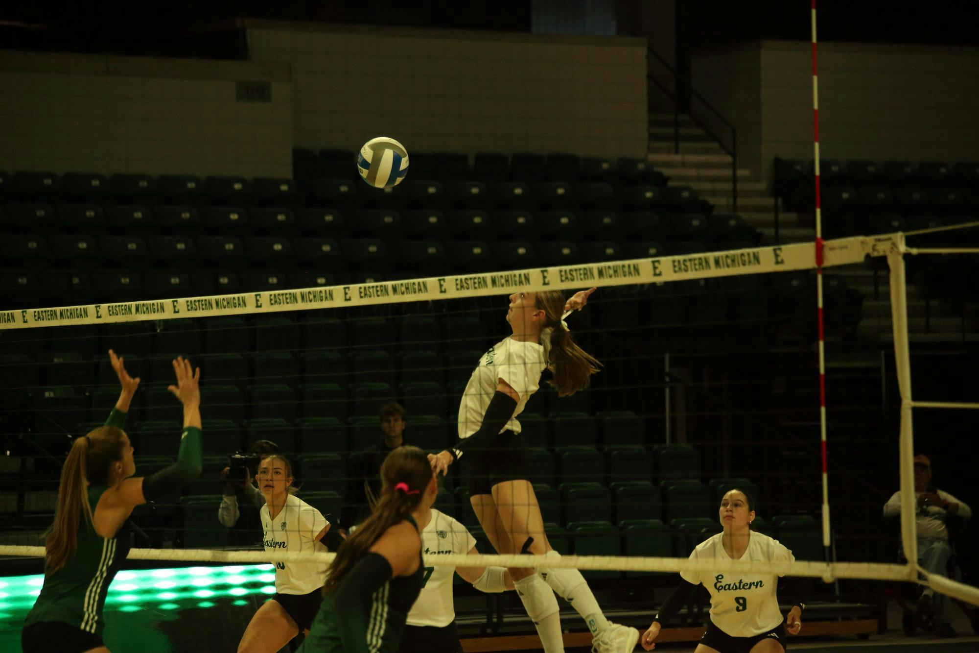 Mid-air women's volleyball player prepares to hit the ball back over the net while teammates watch from the background.