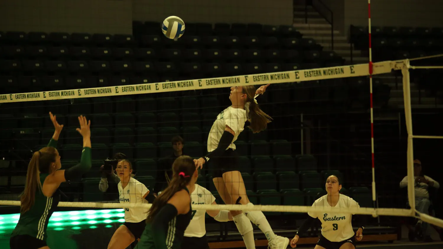 Mid-air women's volleyball player prepares to hit the ball back over the net while teammates watch from the background.