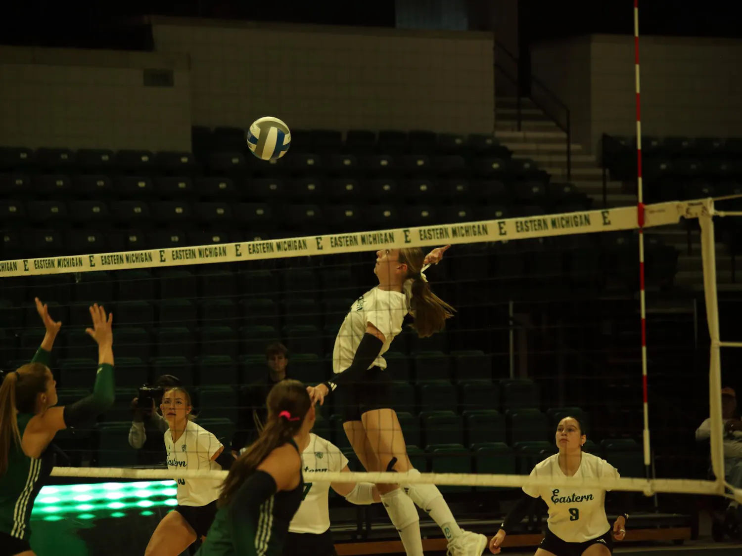 Mid-air women's volleyball player prepares to hit the ball back over the net while teammates watch from the background.