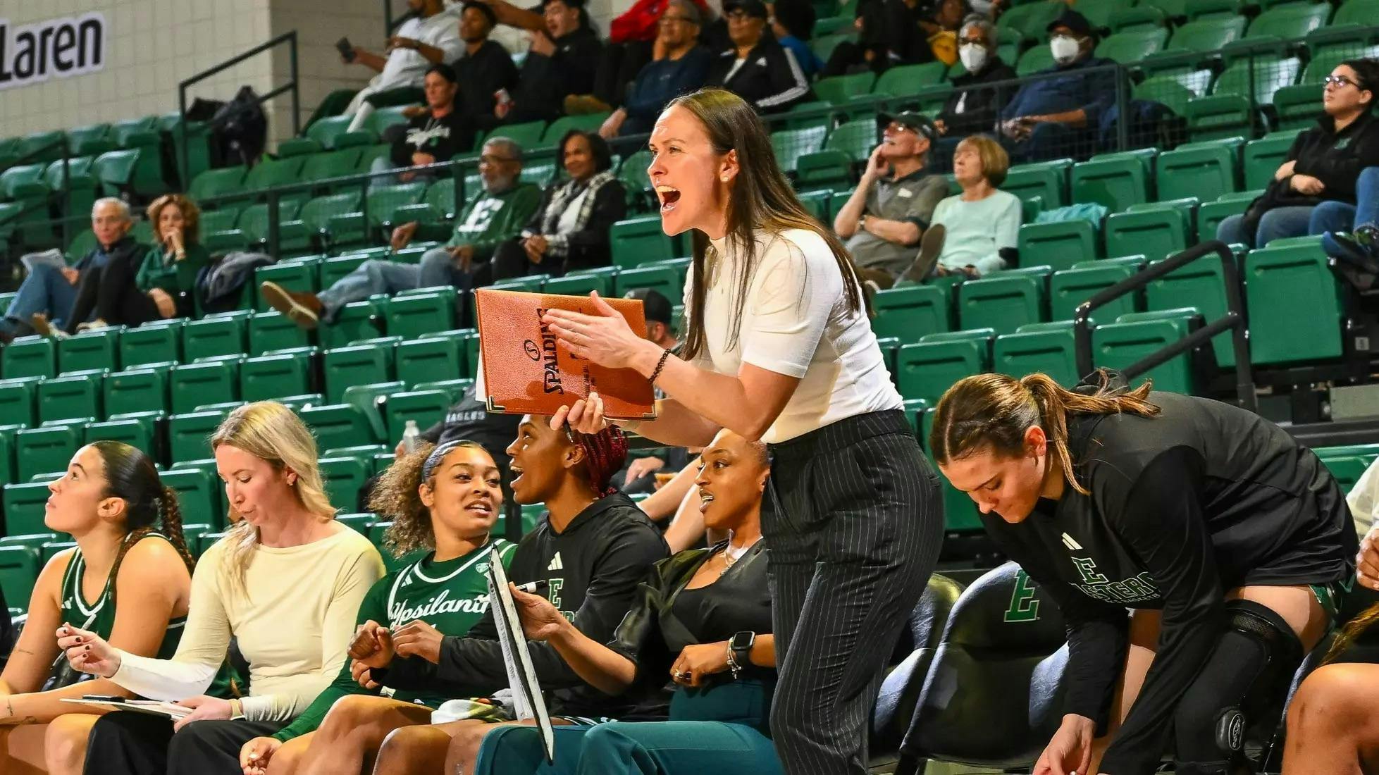 Coach Megan Cunningham smiles, claps, and cheers in front of the bleachers while holding an orange, basketball-colored folder that reads "Spalding." Players and onlookers sit in the bleachers behind her.