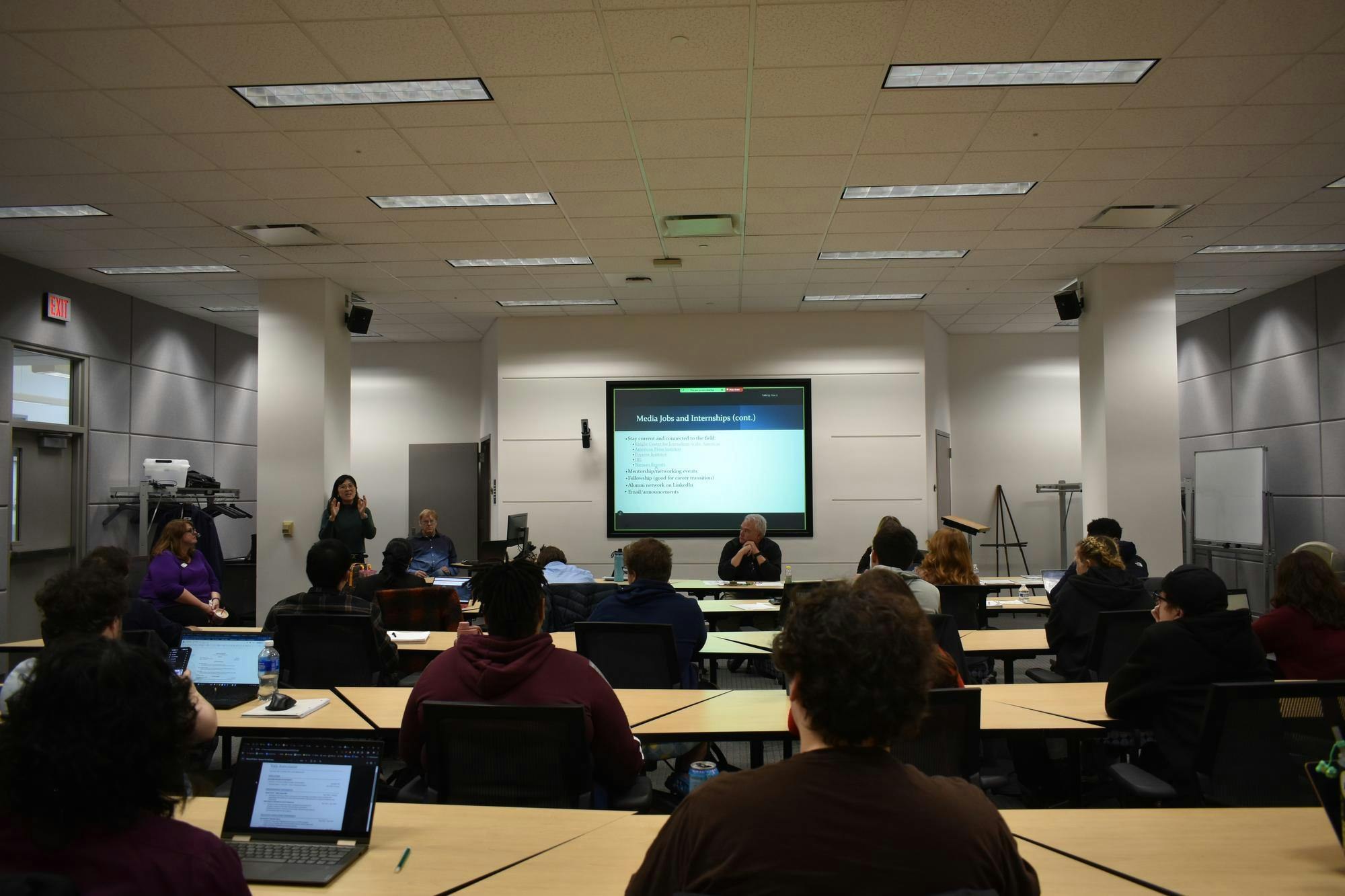 Professor You Li stands at the front of the room with other professors while rows of students sit behind tables and take notes. 
