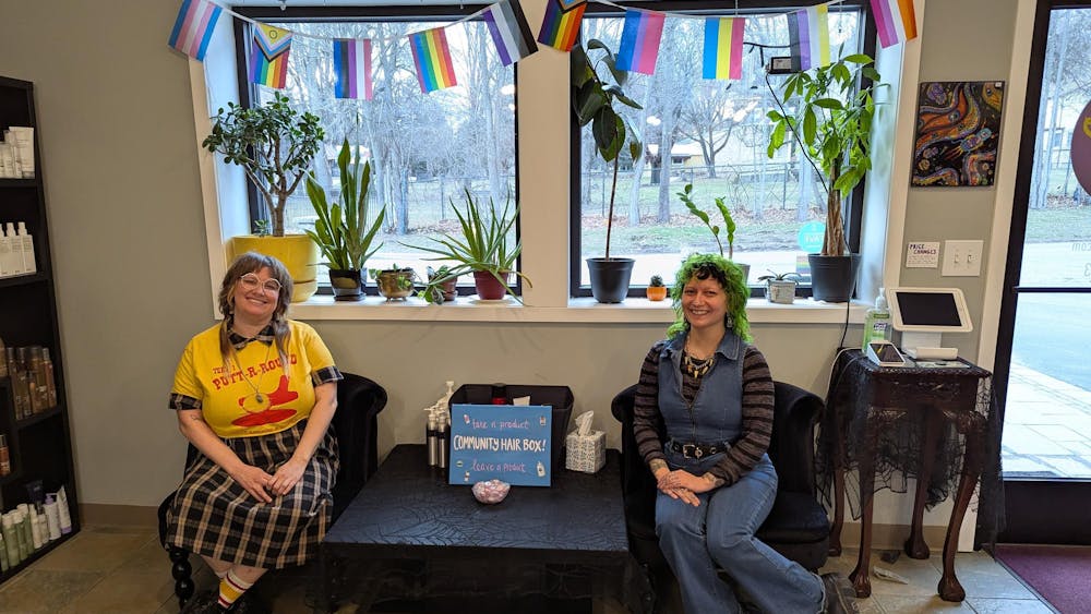 Two people sitting in chairs with pride flags behind them.