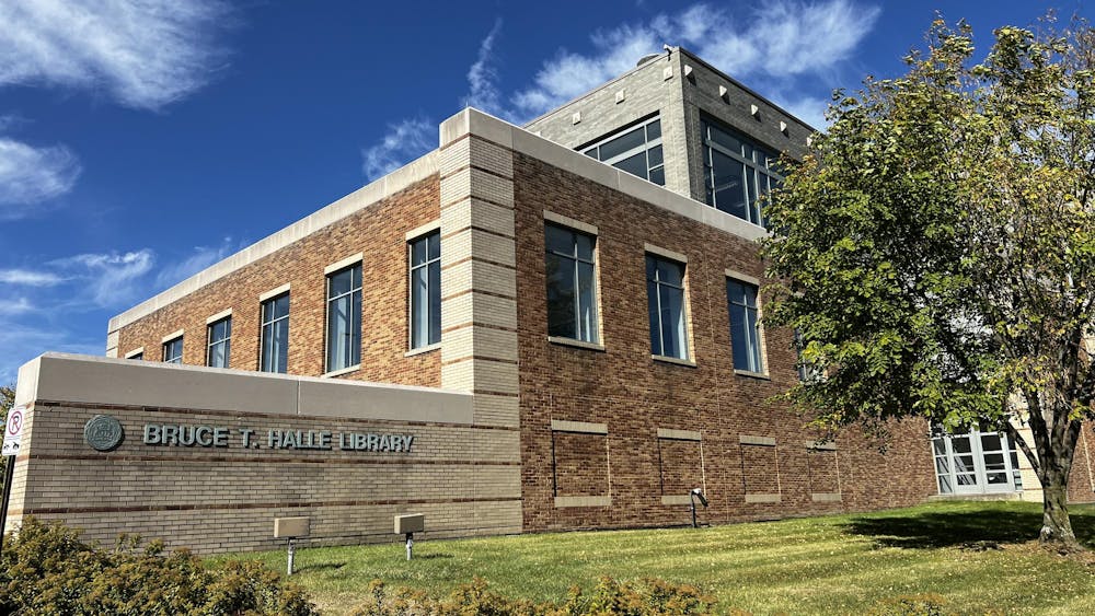 A large brick library building with multiple floors. Letters on the front read "Bruce T. Halle Library." It is a sunny day, and the sky above the building is blue with a few white clouds.