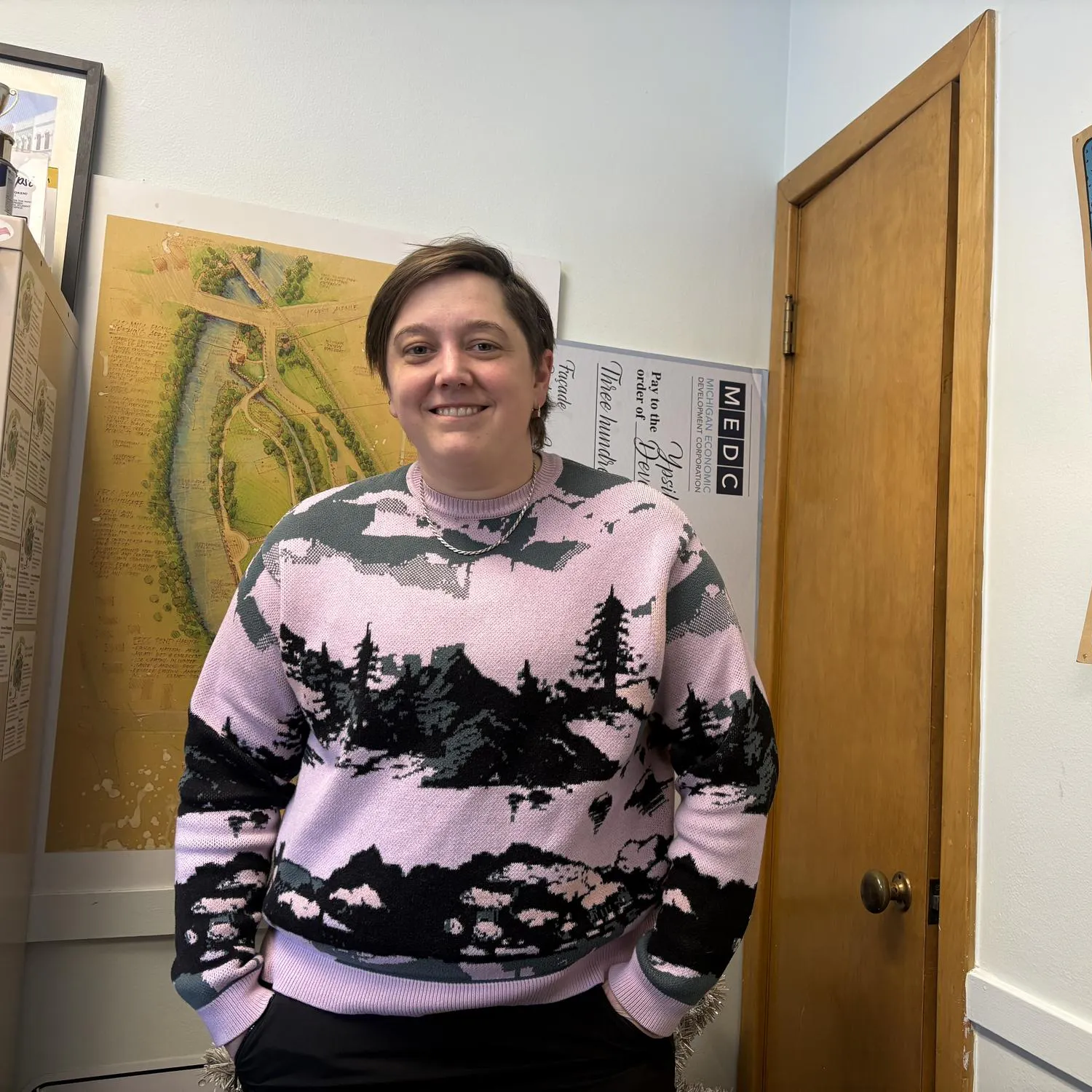 Eliza Jekabson stands in an office smiling; in the background there are colorful posters.