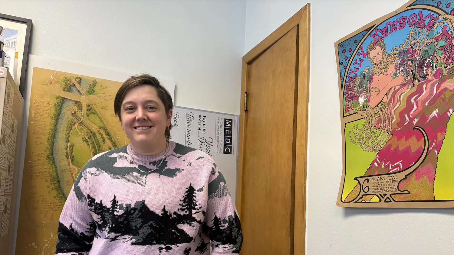 Eliza Jekabson stands in an office smiling; in the background there are colorful posters.