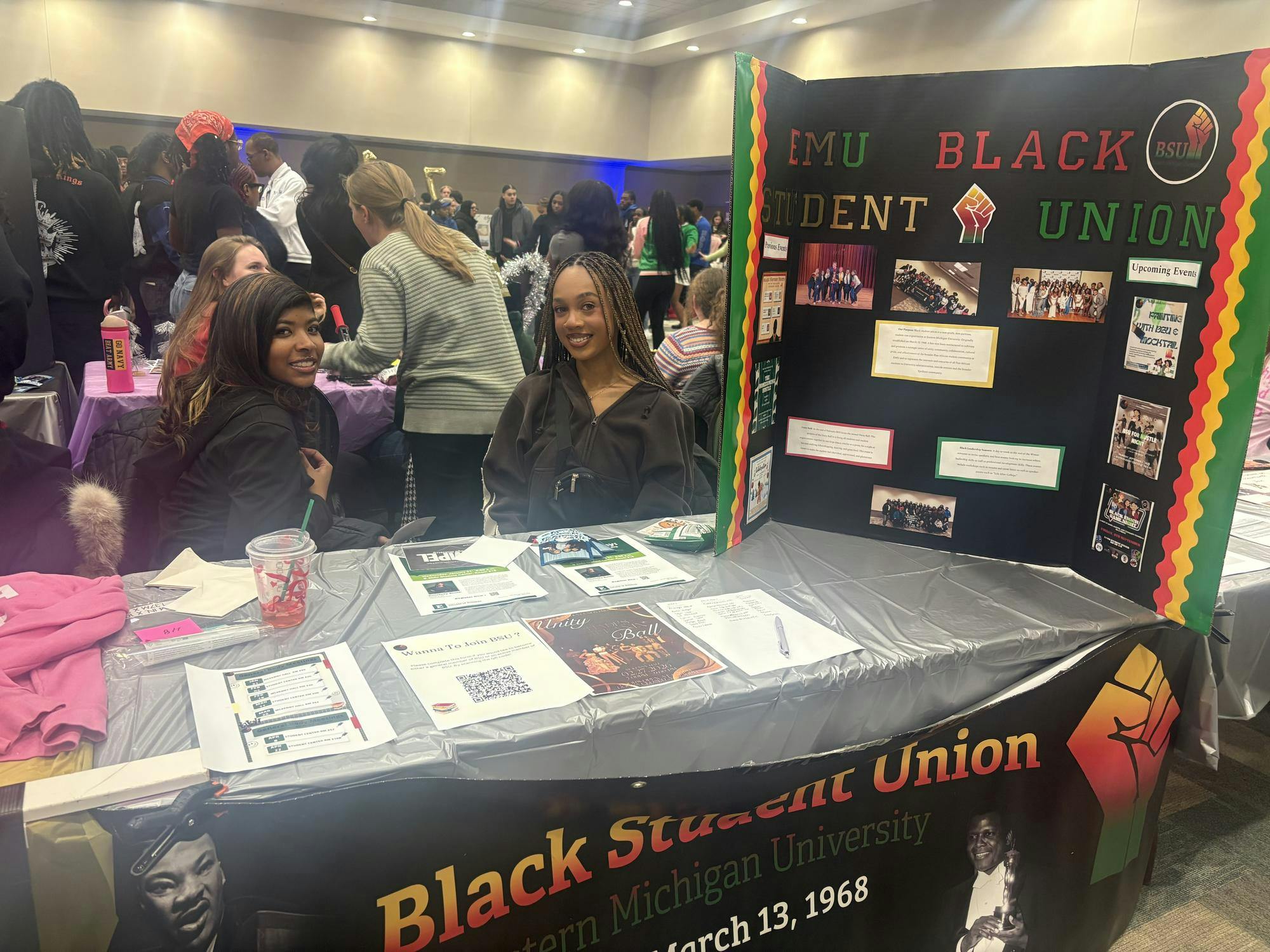 Two women sit behind a table featuring flyers and a large, tri-folding poster for the EMU Black Student Union. Many people gather in the background.