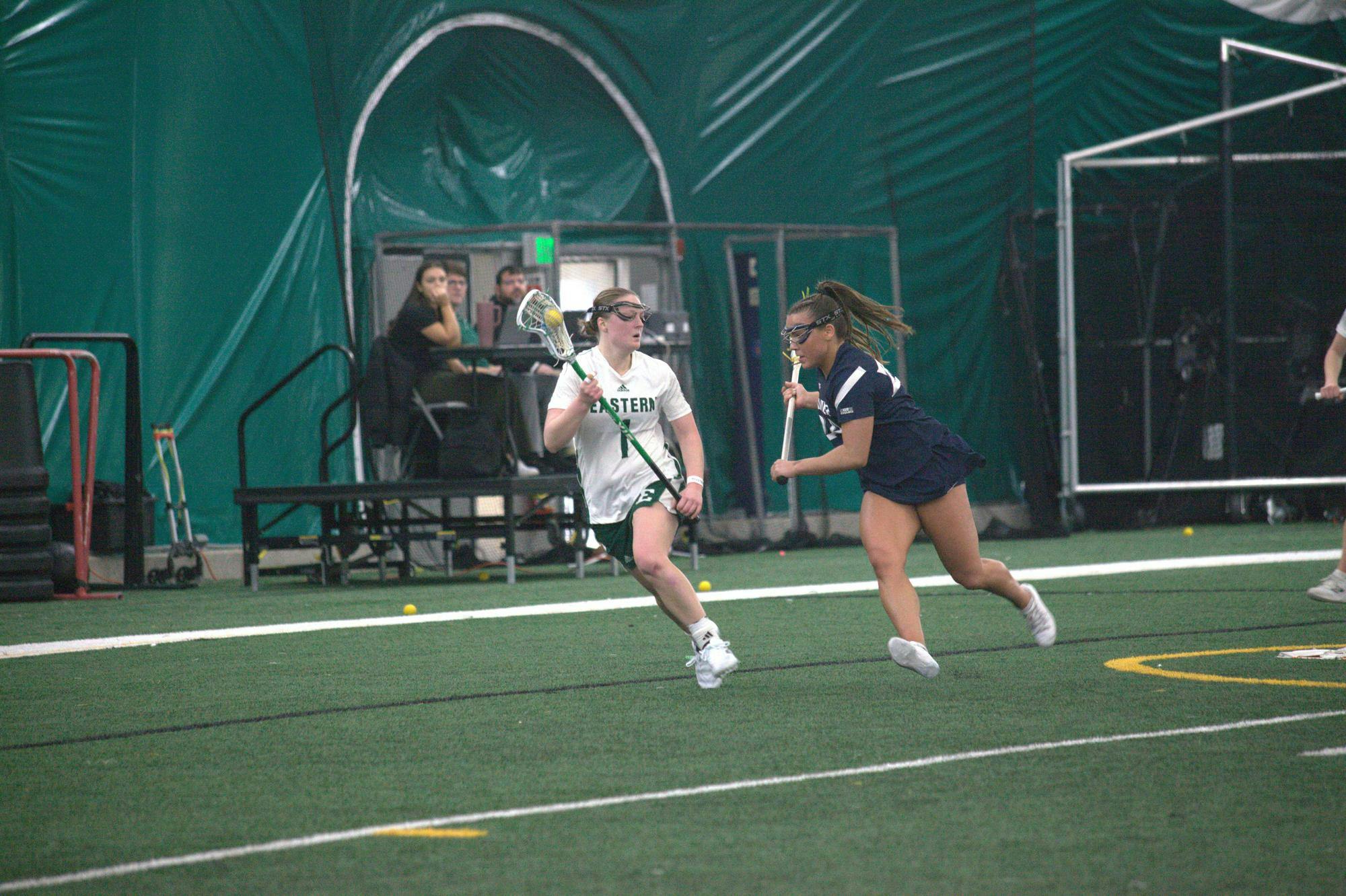 Women's lacrosse player with ball in the pocket of their stick dodges out of the way of opposing team player on the field.