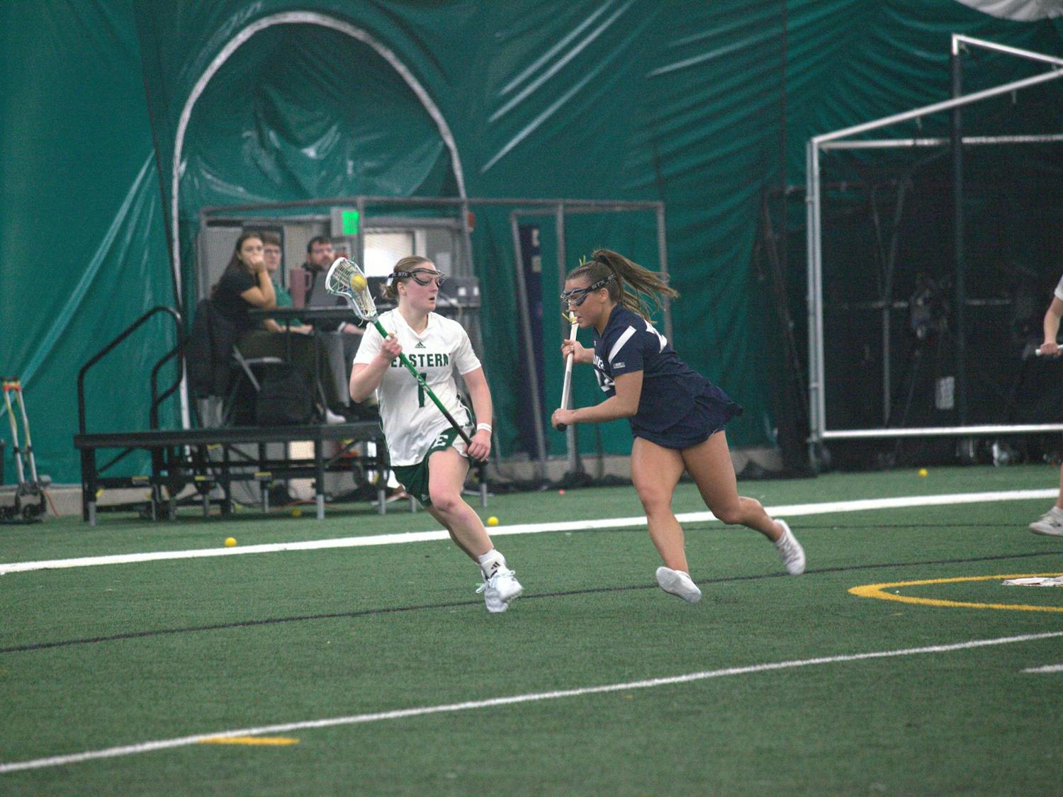 Women's lacrosse player with ball in the pocket of their stick dodges out of the way of opposing team player on the field.