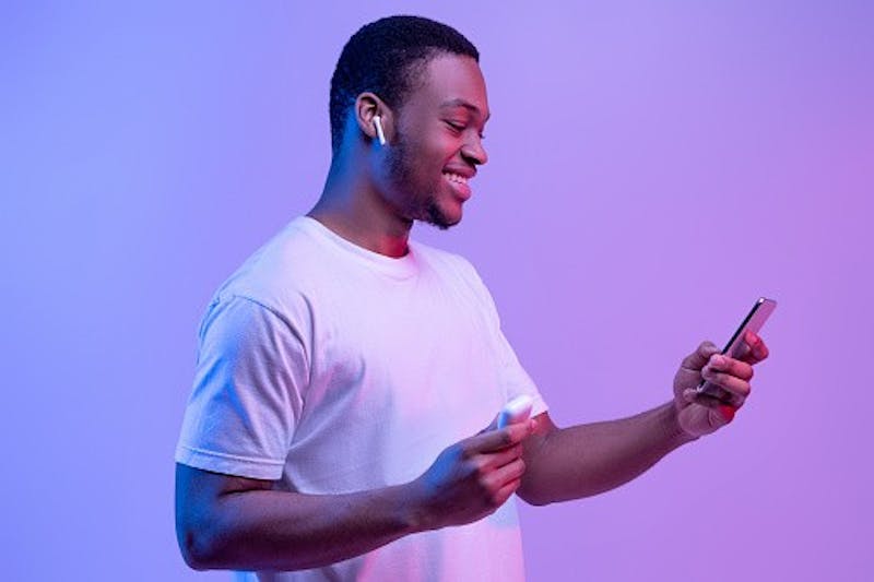 Tech Gadgets For Entertainment. Happy Black Guy Using Smartphone And Earpods For Listening Music, Holding Earphones Case In Hand, Cheerful African Man Standing In Neon Light Over Purple Background