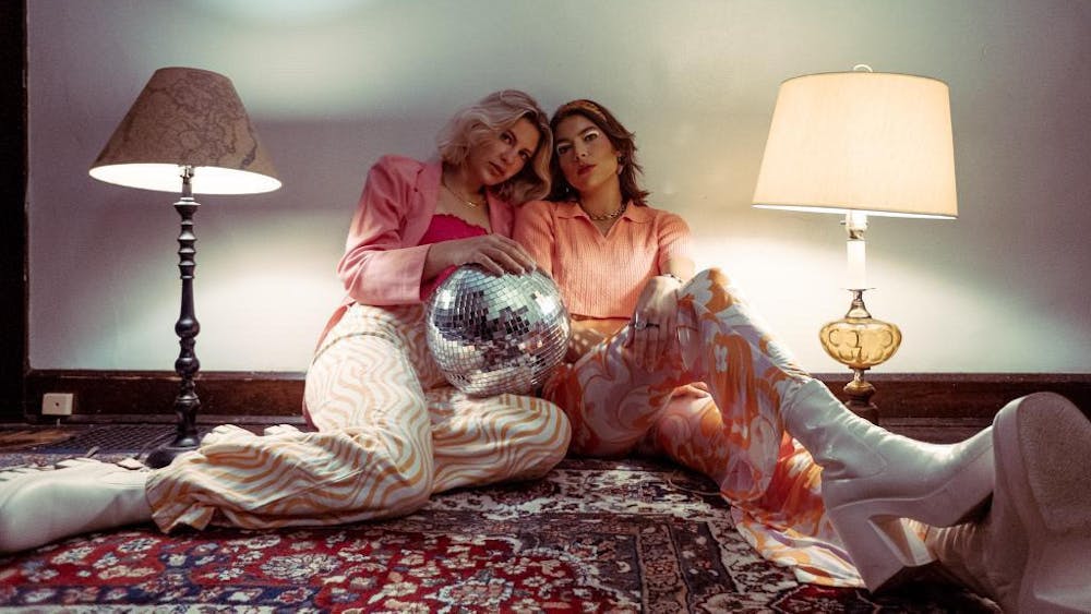 Julia Rusyniak and Emma Spartz pose between two lamps with a disco ball in their laps. The two women are lead vocalists in the band Six Foot Blonde.