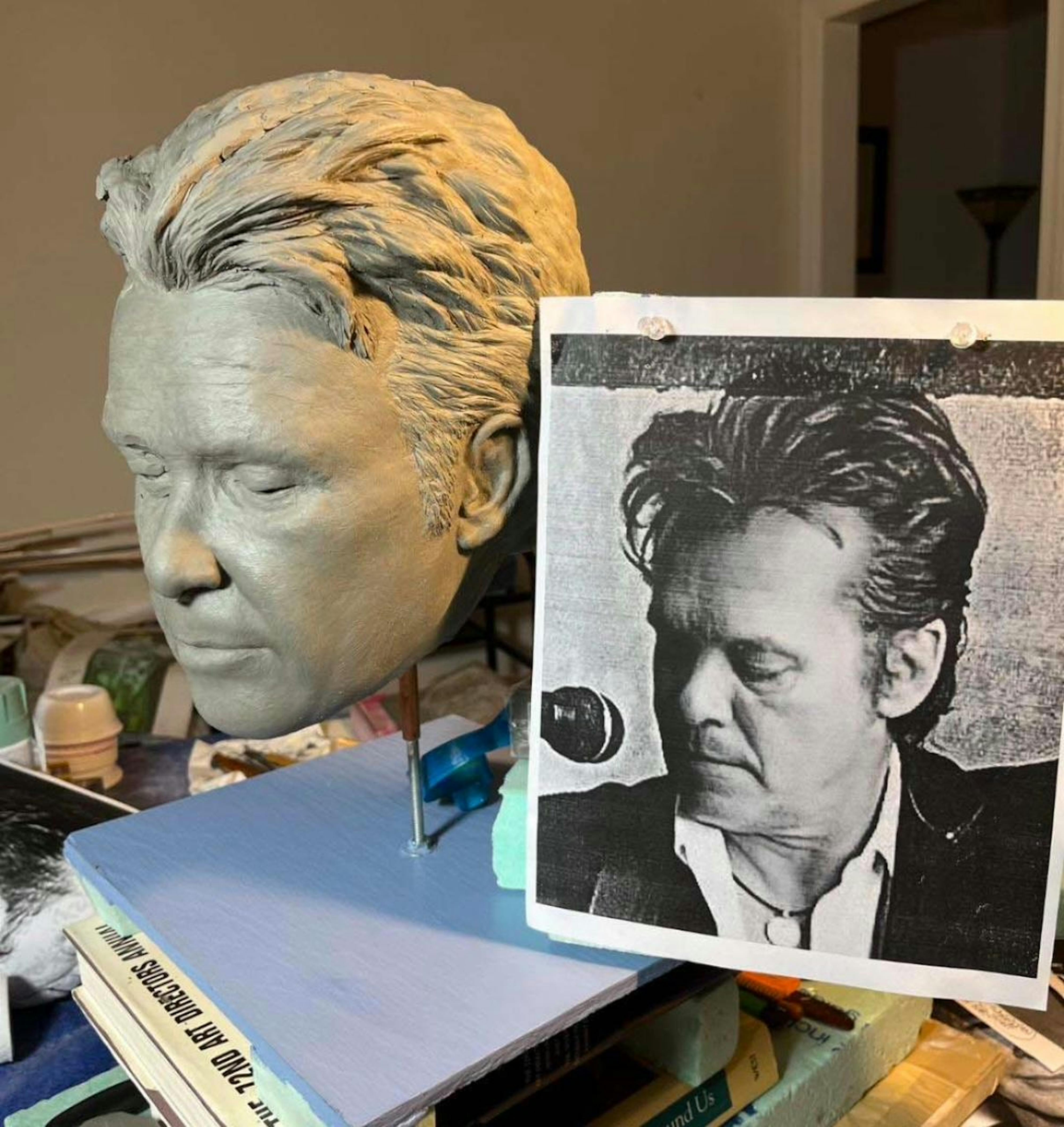 A gray clay sculpture of a man's face with closed eyes sits on a desk next to a printed black-and-white photo of the same face.