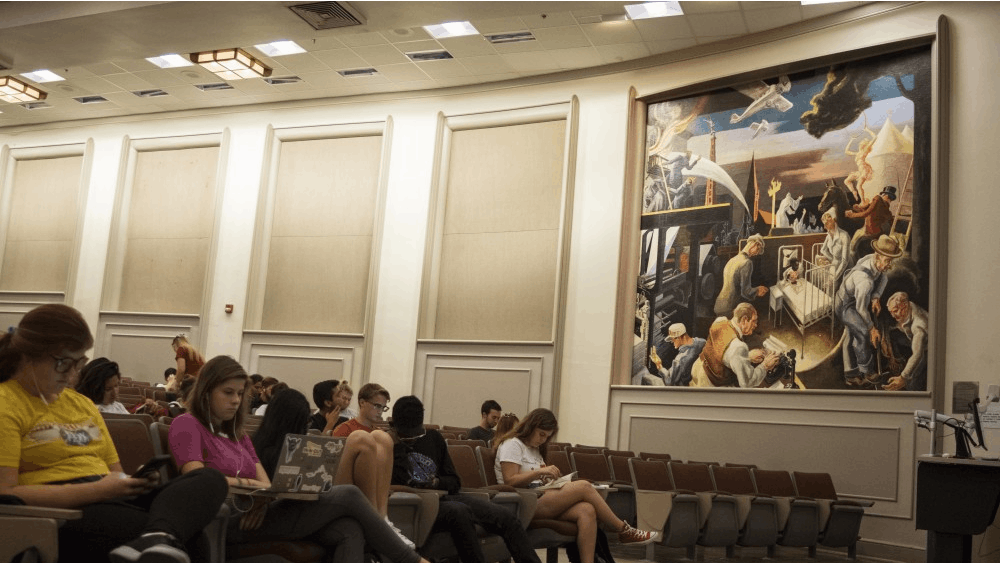 Students wait for class to begin in Woodburn 100. The lecture hall contains a mural created by Thomas Hart Benton in 1933, which has created controversy for its depiction of hooded Ku Klux Klan members in its background.&nbsp;