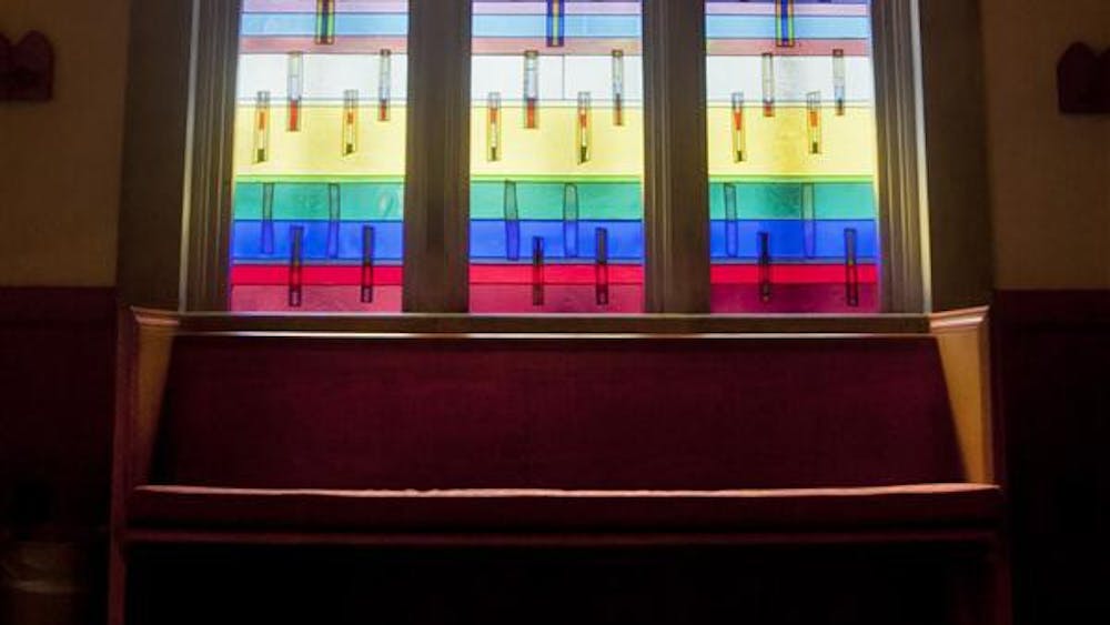 The sun shines through stained glass behind a pew. 