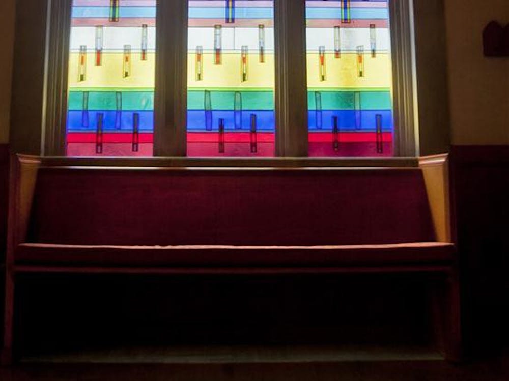 The sun shines through stained glass behind a pew. 
