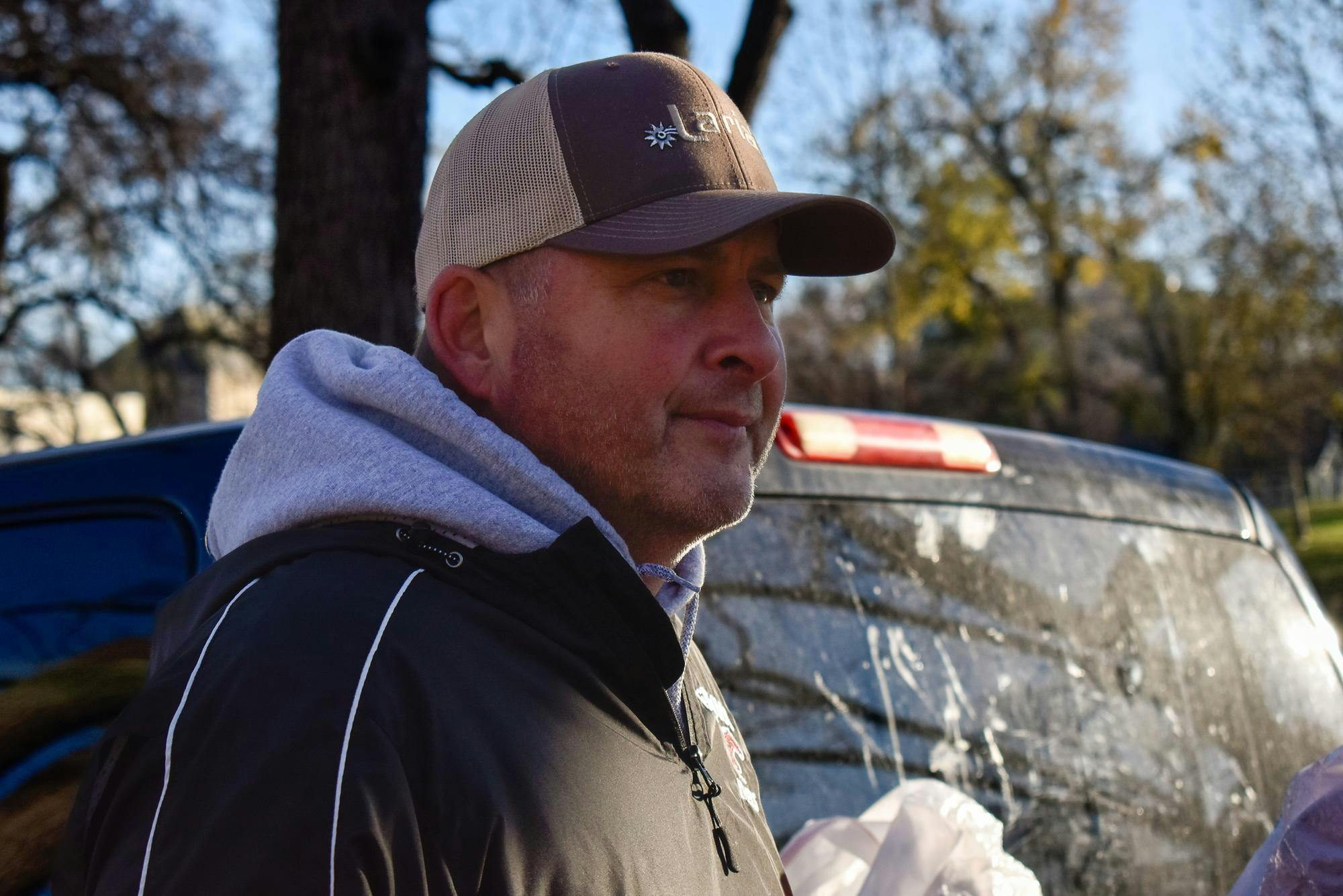 A man in a baseball cap and hoodie stands in front of his truck looking to the right.