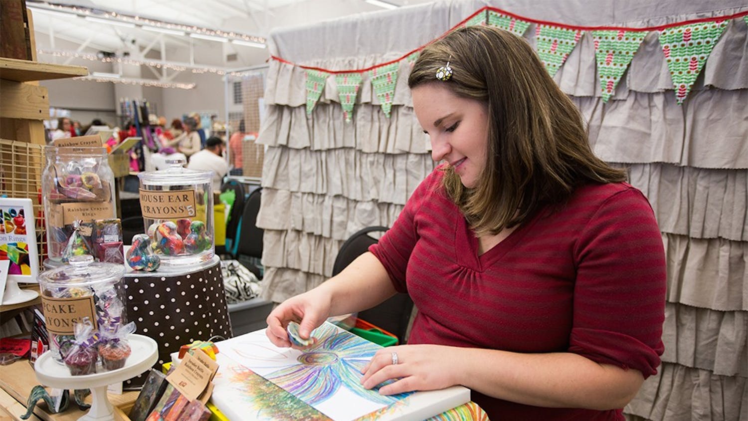 Artist Nicole Lewis draws rainbow flowers with her handmade rainbow crayon at the Bloomington Handmade Market on Nov. 8, 2014. "Art 2 the Extreme" features recycled, rainbow crayon creations in over 175 shapes and sizes. Teacher by day and artist by night, Nicole creates fun and unique gifts for all ages and interests. From crayon rings to giant, crayon robots, there is something for everyone to enjoy in their stocking this holiday." ( Bloomington Handmade Market Guide Book, 47)
