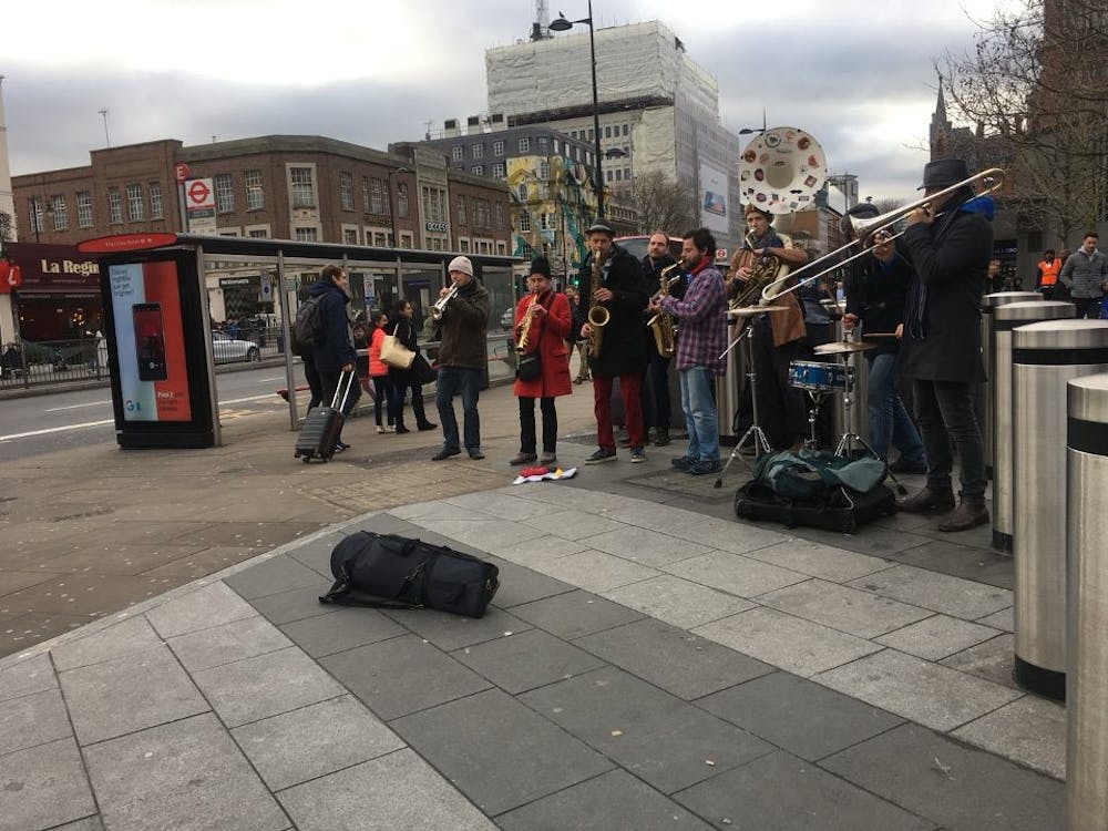A group of seven musicians stands outside the King’s Cross tube station playing instruments. The group is one of many that can be seen on the streets of London.