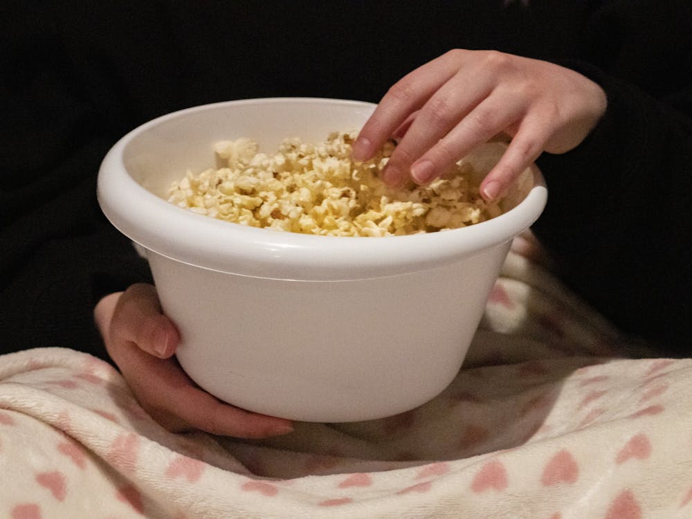 A students holds a bowl of popcorn while sitting on the couch watching a movie. 