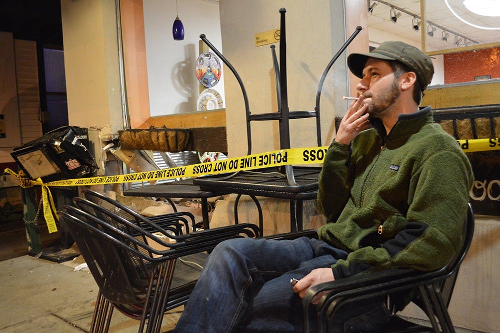 Tyler Brown smokes a cigarette in the chair he was sitting in when he witnessed a car crashed into the Bloomington Bagel Company Wednesday evening.