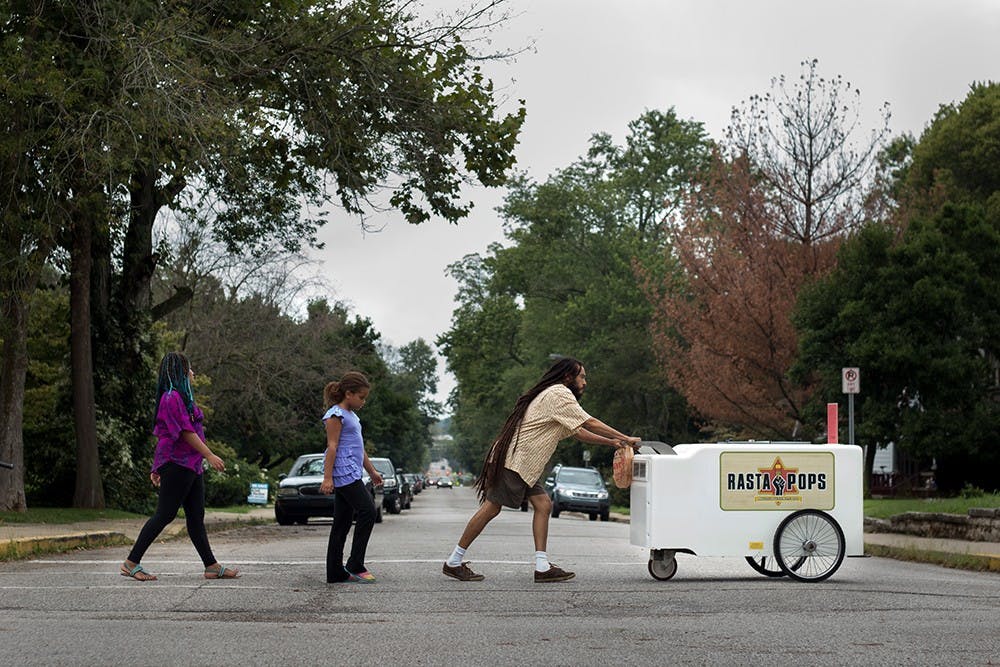 Iuri Santos pushes the Rasta Pops cart toward downtown Bloomington with his daughter Zara (left), 10, and her friend Jasmine following behind. Santos grew up in Brazil where he says vendors are on almost every street selling ice pops from coolers strapped to their necks, it was those memories that inspired him and his wife to start this business in Bloomington. 