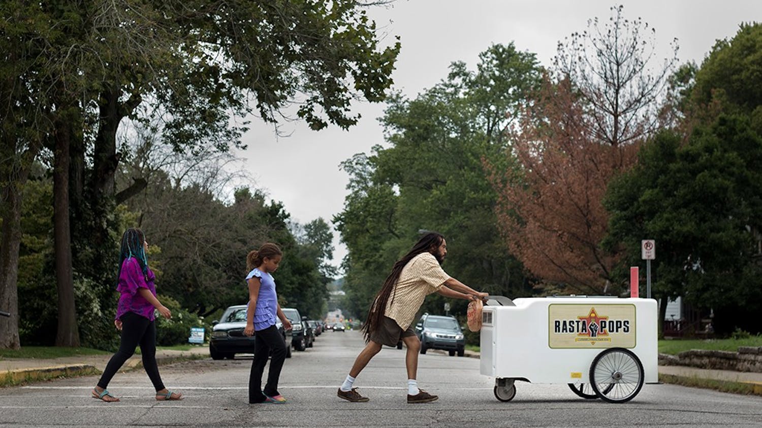 Iuri Santos pushes the Rasta Pops cart toward downtown Bloomington with his daughter Zara (left), 10, and her friend Jasmine following behind. Santos grew up in Brazil where he says vendors are on almost every street selling ice pops from coolers strapped to their necks, it was those memories that inspired him and his wife to start this business in Bloomington.