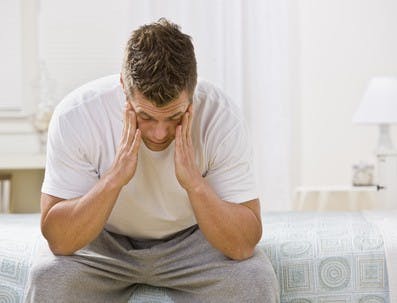 A man is seated on the bed in a bedroom.  He has his head in his hands and is looking away from the camera.  Horizontally framed shot.