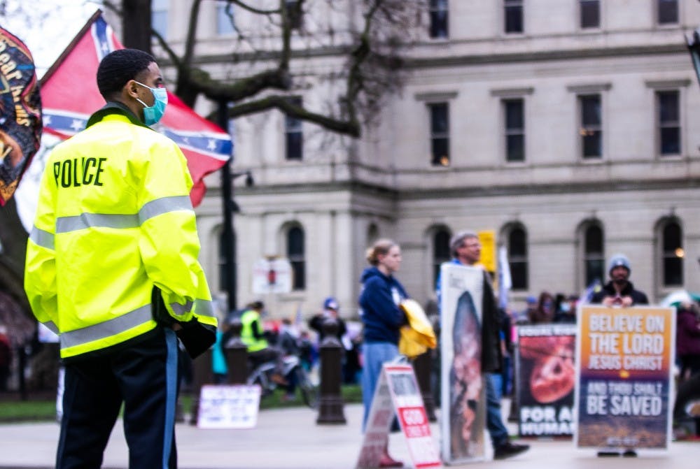 <p>A police officer in a mask stands directing traffic as a truck with a confederate flag drives past the rally at the Capitol April 30, 2020 in Lansing. Crowds gathered to protest Governor Whitmer&#x27;s stay-at-home order. The protest started outside before crowds moved inside where they were stopped from entering the House and Senate due to social distancing guidelines. While the protest was namely about the stay-at-home order, individual protestors&#x27; priorities varied from abortion to President Trump&#x27;s reelection.</p>