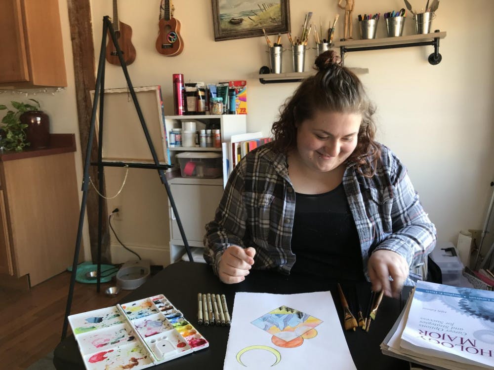Emily Brewer sits with her water colors and paint brushes explaining her technique for her illustrations. The piece in front is titled, "The Pass" themed by the poem of the same name. 
