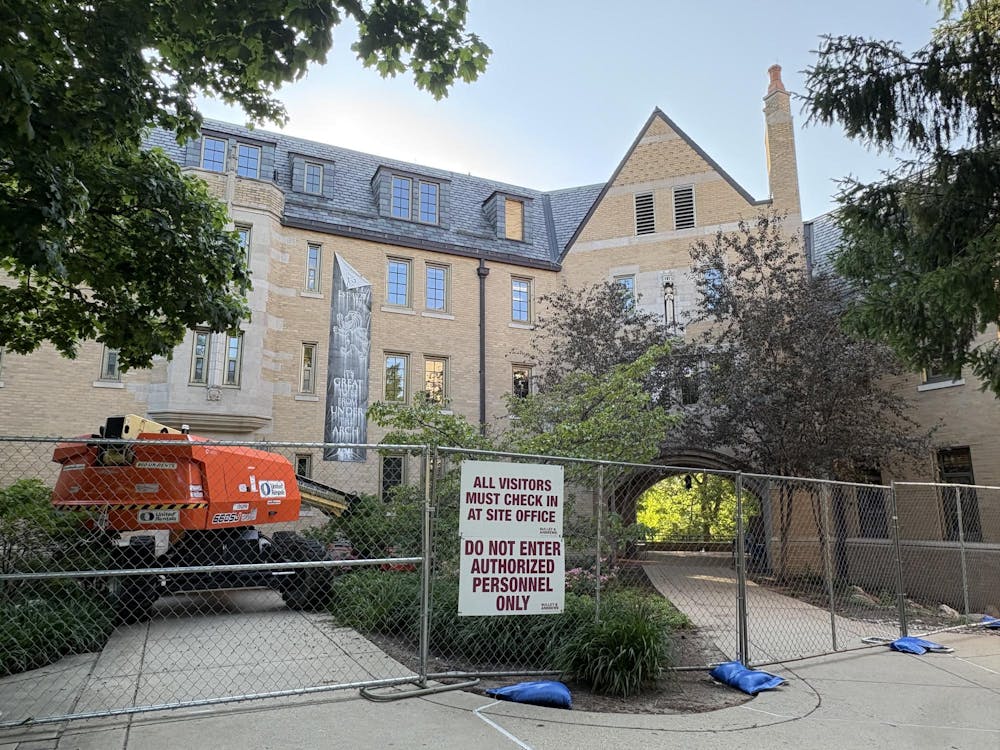 Lyons Hall, a beige brick building, has weathered sign hanging from it. Building is surrounded by construction equipment and chain link fence with signs reading "All visitors must check in at site office" and "Do not enter authorized personnel only."