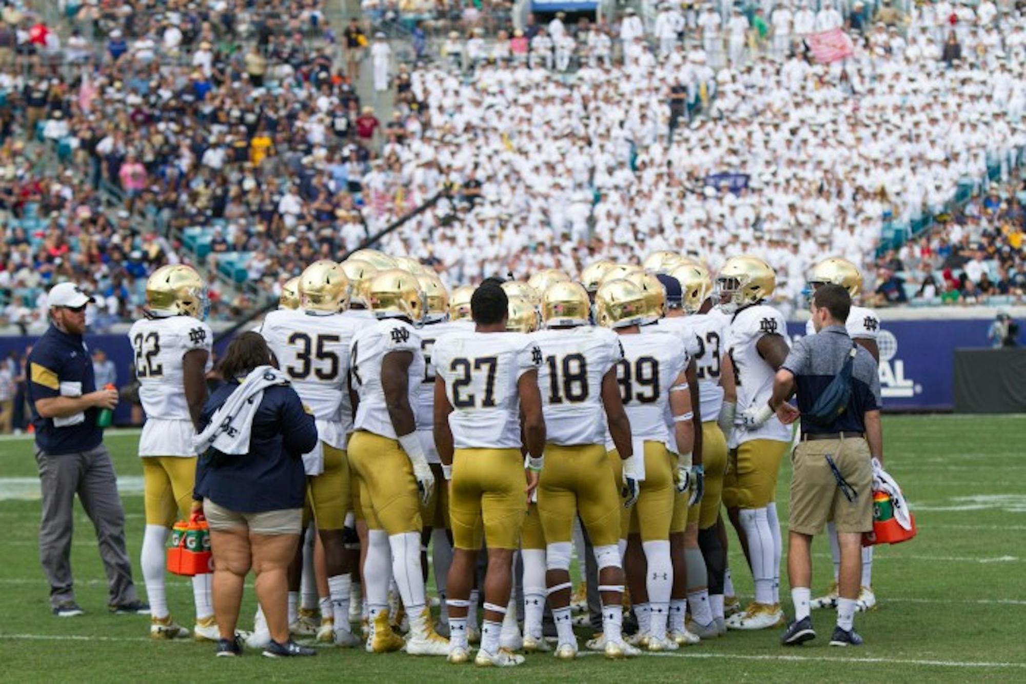 Notre Dame meets in a huddle during 4th quarter. The midshipmen stand cheering in their formal uniforms in the background.