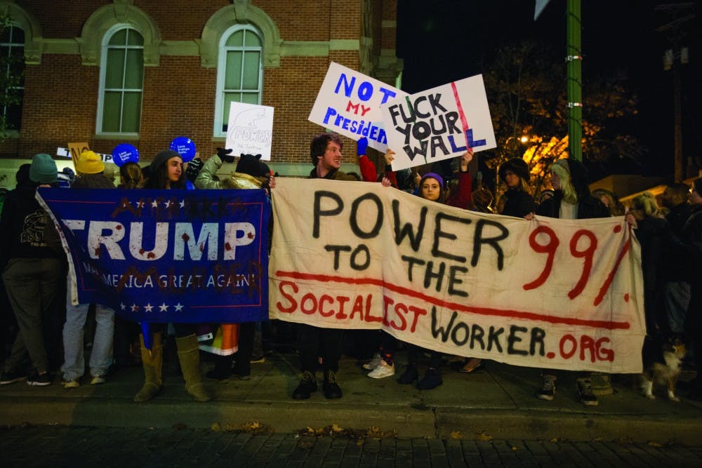 Natalie Mahomar (holding the Trump/Pence sign), Grant Stover and Alana Baldwin (holding the Socialist Worker sign), and Emily Goldberg (holding the anti-wall sign) protest outside of the Athens County Courthouse on Sunday, November 13. Mahomar's Trump flag says "AmeriKKKa, Trump equals murder." 