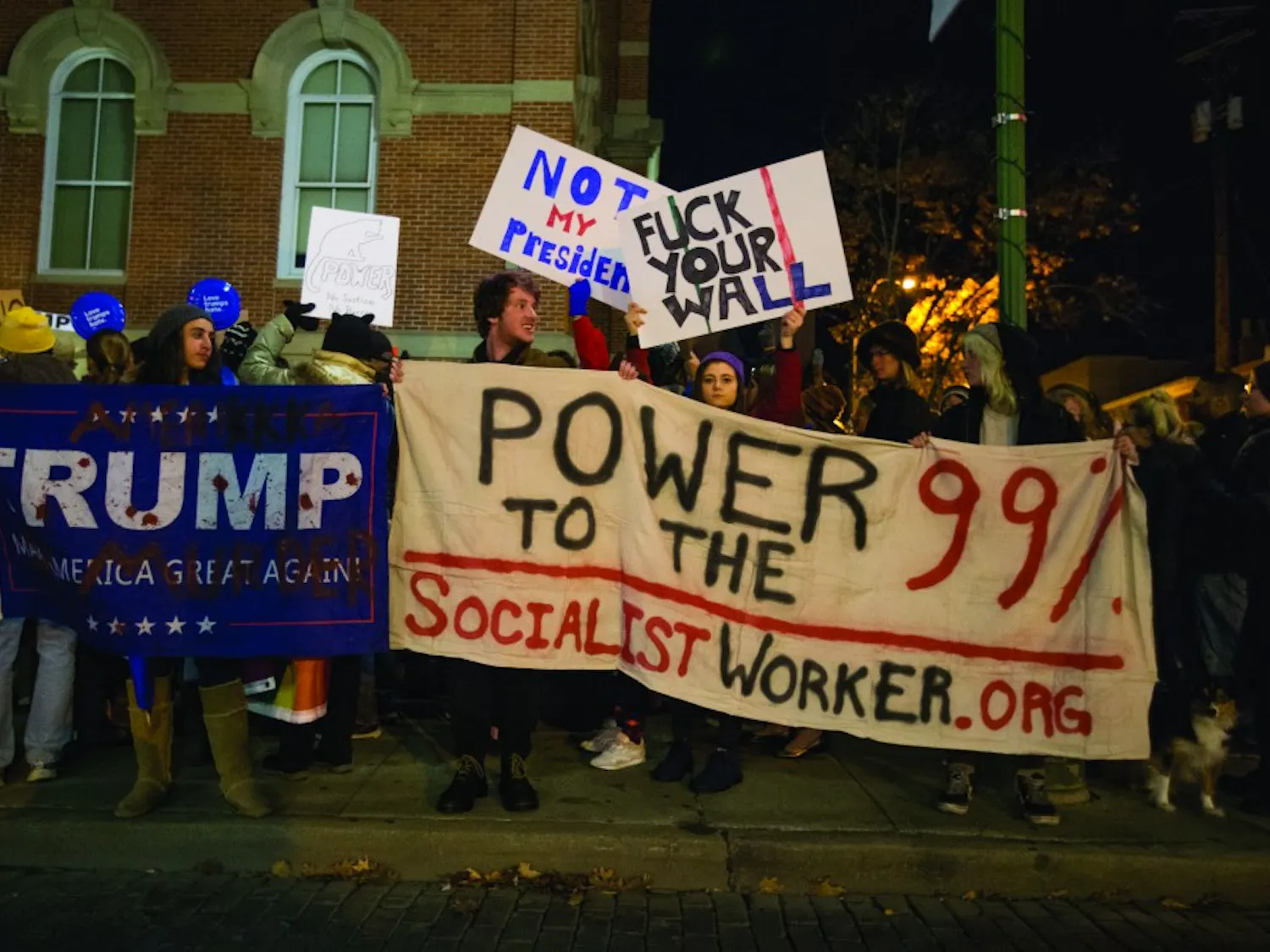 Natalie Mahomar (holding the Trump/Pence sign), Grant Stover and Alana Baldwin (holding the Socialist Worker sign), and Emily Goldberg (holding the anti-wall sign) protest outside of the Athens County Courthouse on Sunday, November 13. Mahomar's Trump flag says "AmeriKKKa, Trump equals murder."