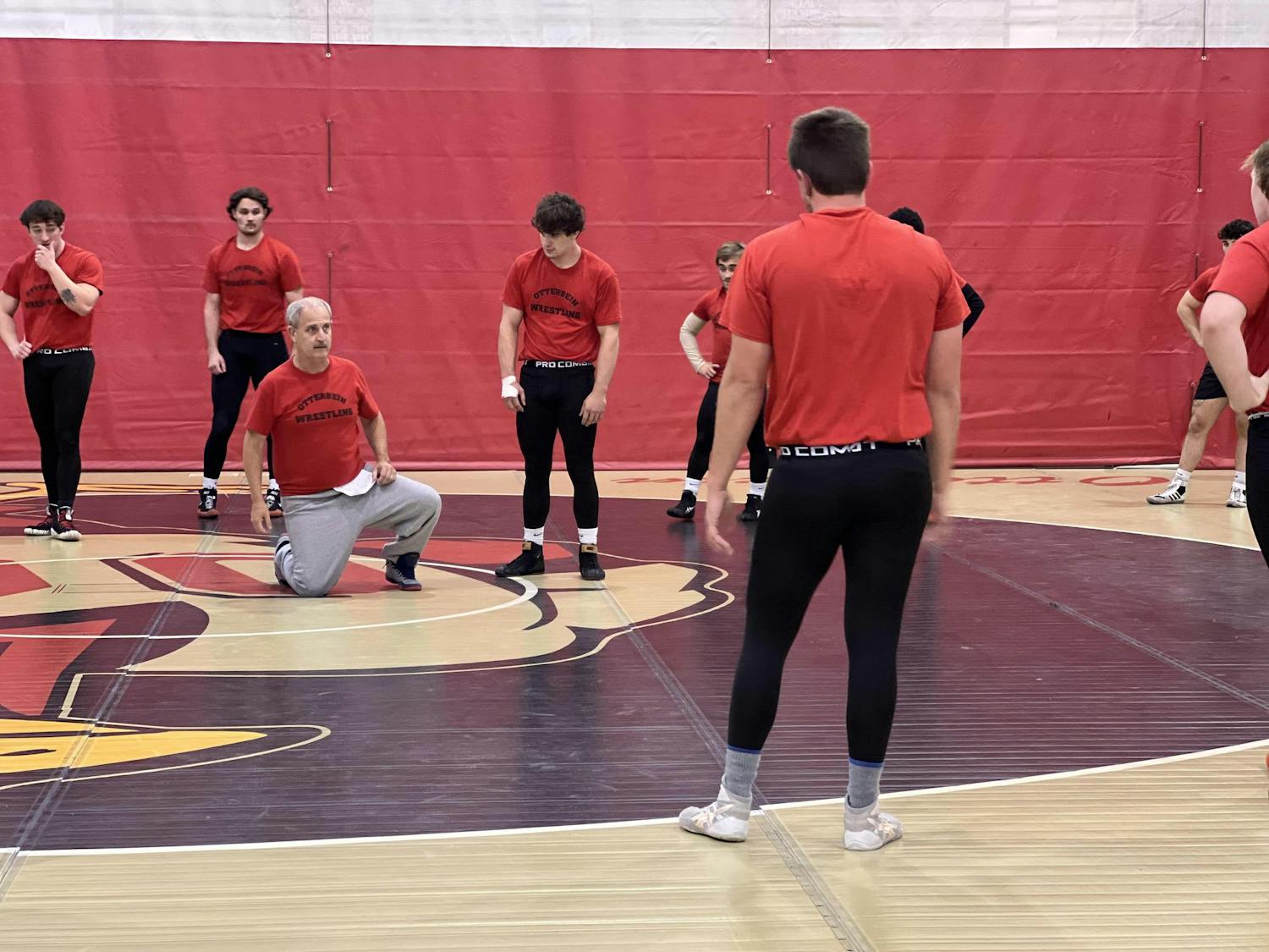 Assistant coach Doug short kneels on a wrestling mat while Otterbein wrestlers watching during a practice session. 