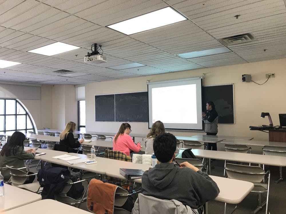 A teacher next to a projector speaks to a classroom of students.