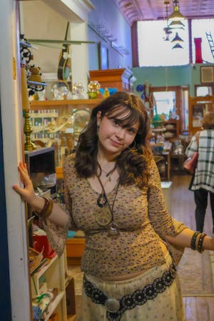 Girl with brown hair wearing necklaces and a brown shirt posing inside of a doorway to an art store.