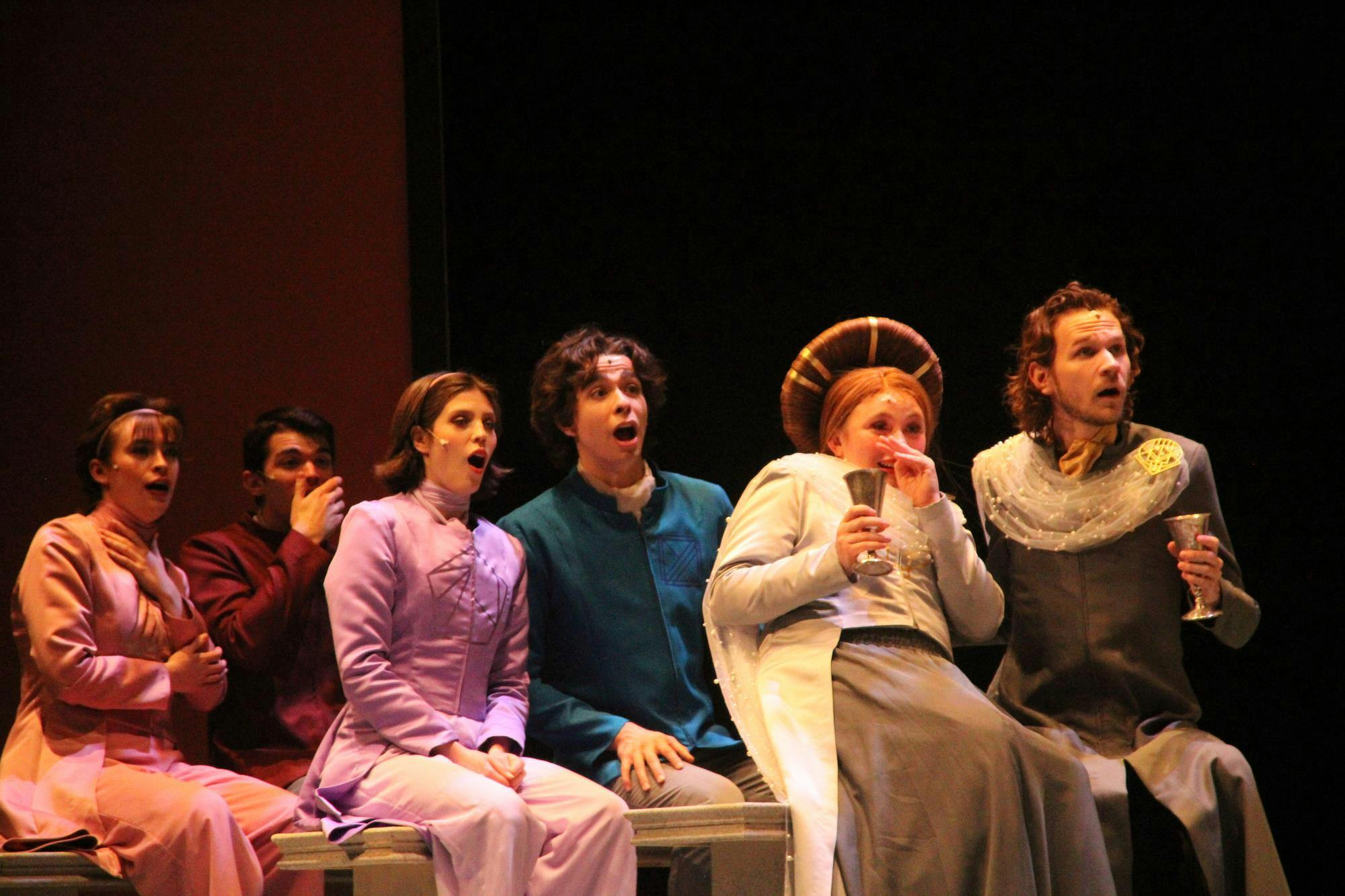 Six Otterbein performers are sitting on benches in full costume, reacting to news they've heard on stage.