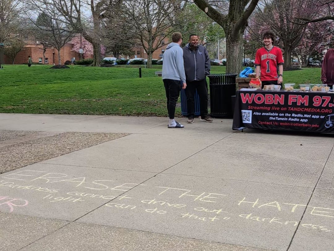 Three people stand near a WOBN table on the library patio. There is chalk on the patio that says "Erase the Hate // Darkness cannot drive out darkness, light can do that."