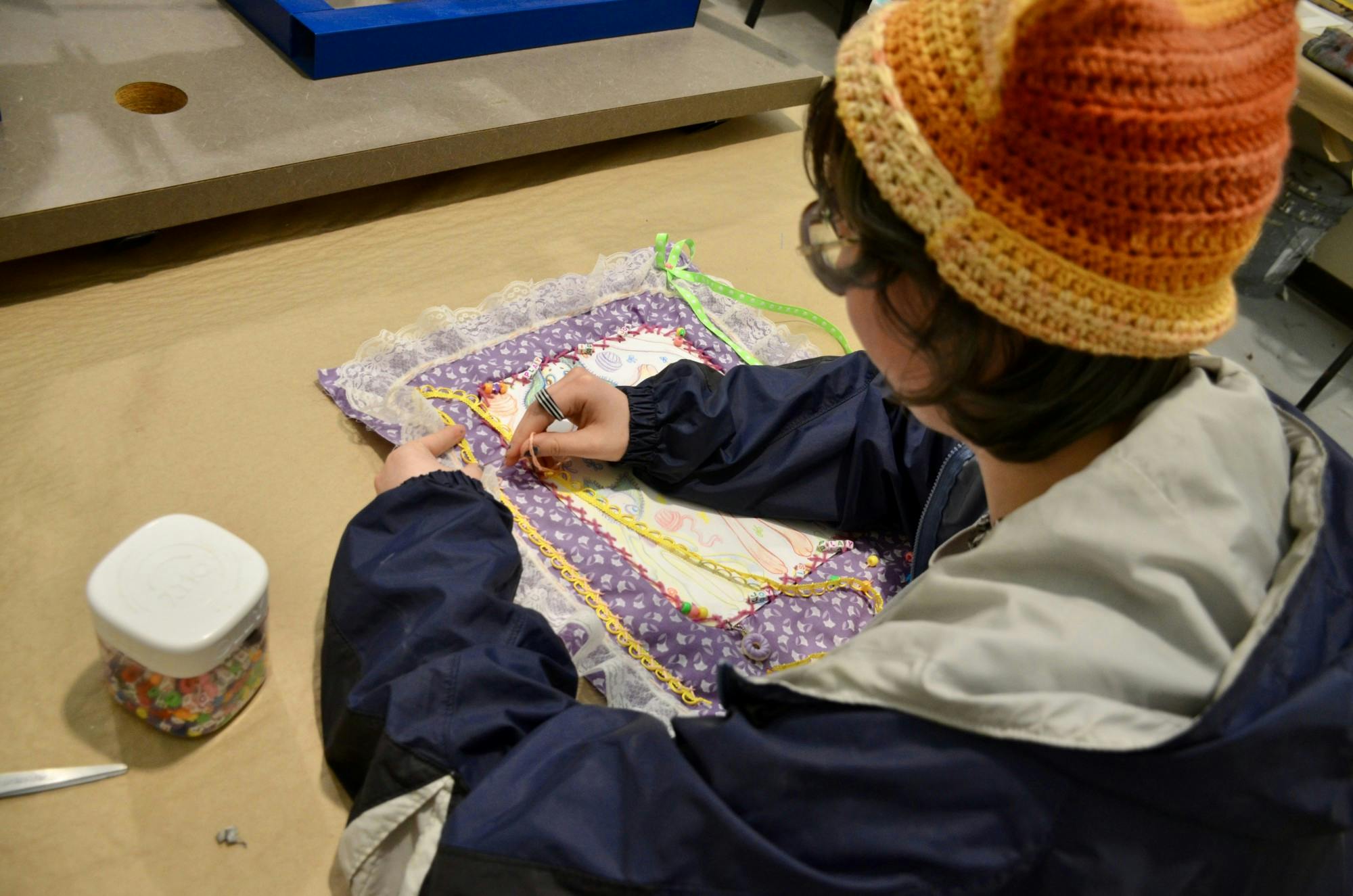 A student works on their senior artwork by sewing elements onto their piece. Their piece is made of purple fabric with lace surrounding it.