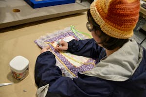 A student works on their senior artwork by sewing elements onto their piece. Their piece is made of purple fabric with lace surrounding it.
