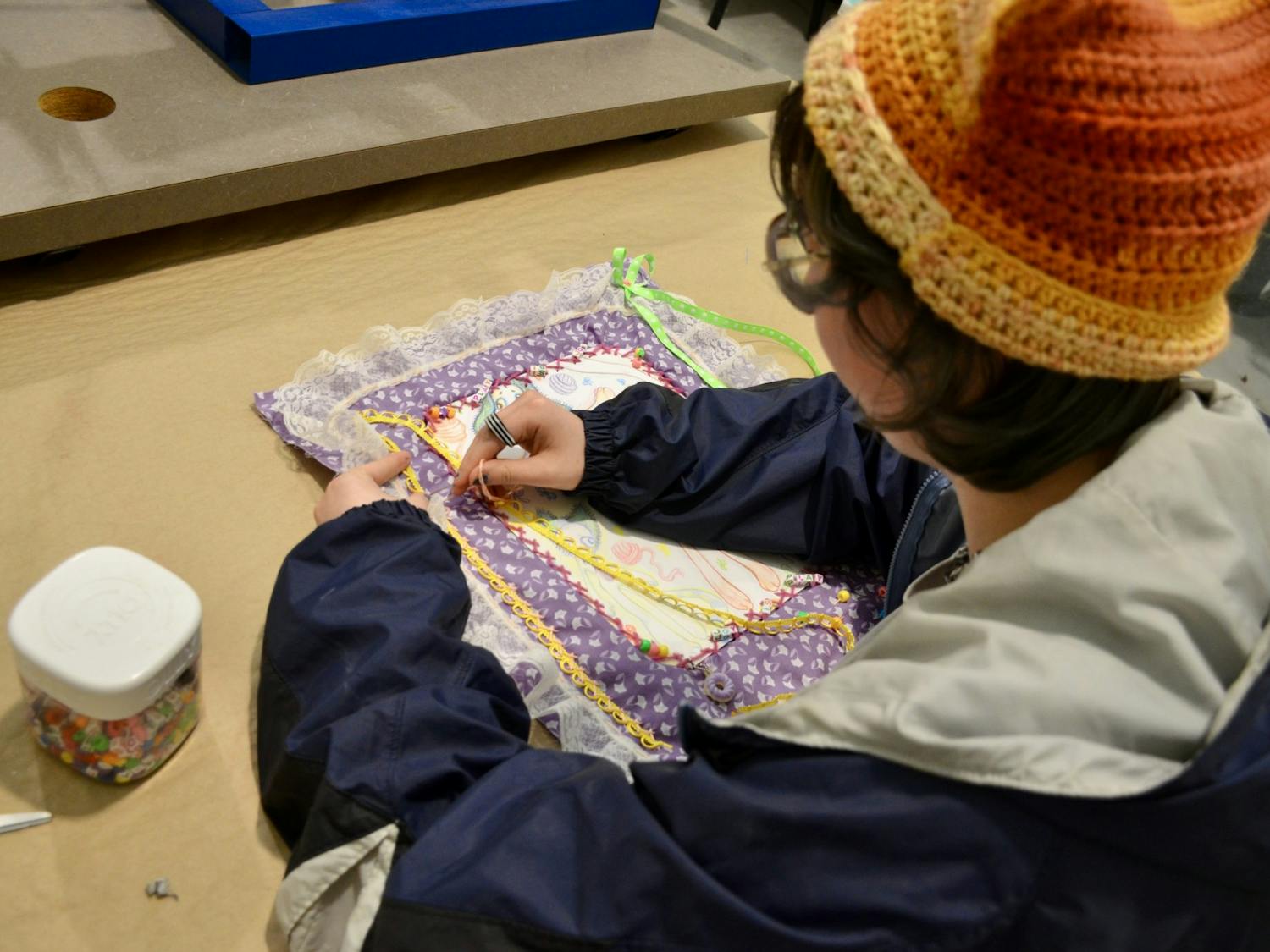 A student works on their senior artwork by sewing elements onto their piece. Their piece is made of purple fabric with lace surrounding it.