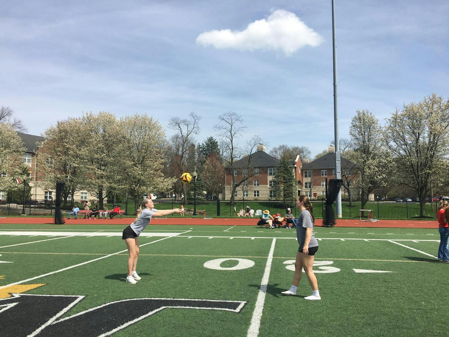 Two girls both in gray shirts and black shorts pass a volleyball between each other on a football field.
