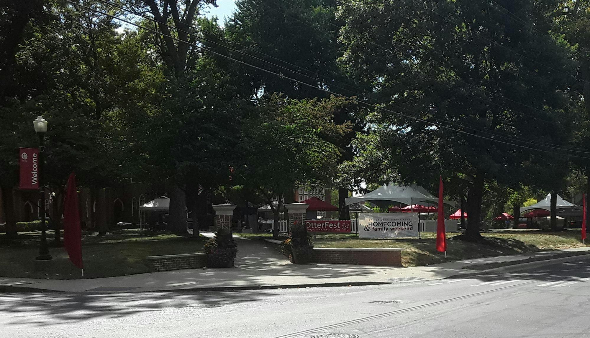 Red and white outdoor tents set up on grass in front of brick building. A red banner reads "OtterFest" and a white banner reads "Homecoming and Family Weekend."