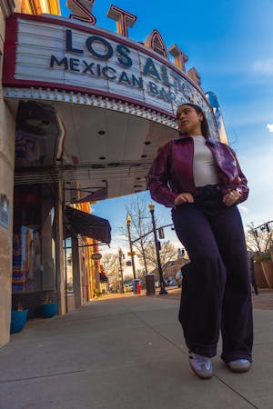 Dark haired girl poses in a purple jacket, jeans, and white shirt posing in front of a restaurant.