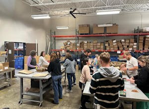 A large family splits between two tables in a warehouse and set up brown paper bags and plastic wrap for food. 