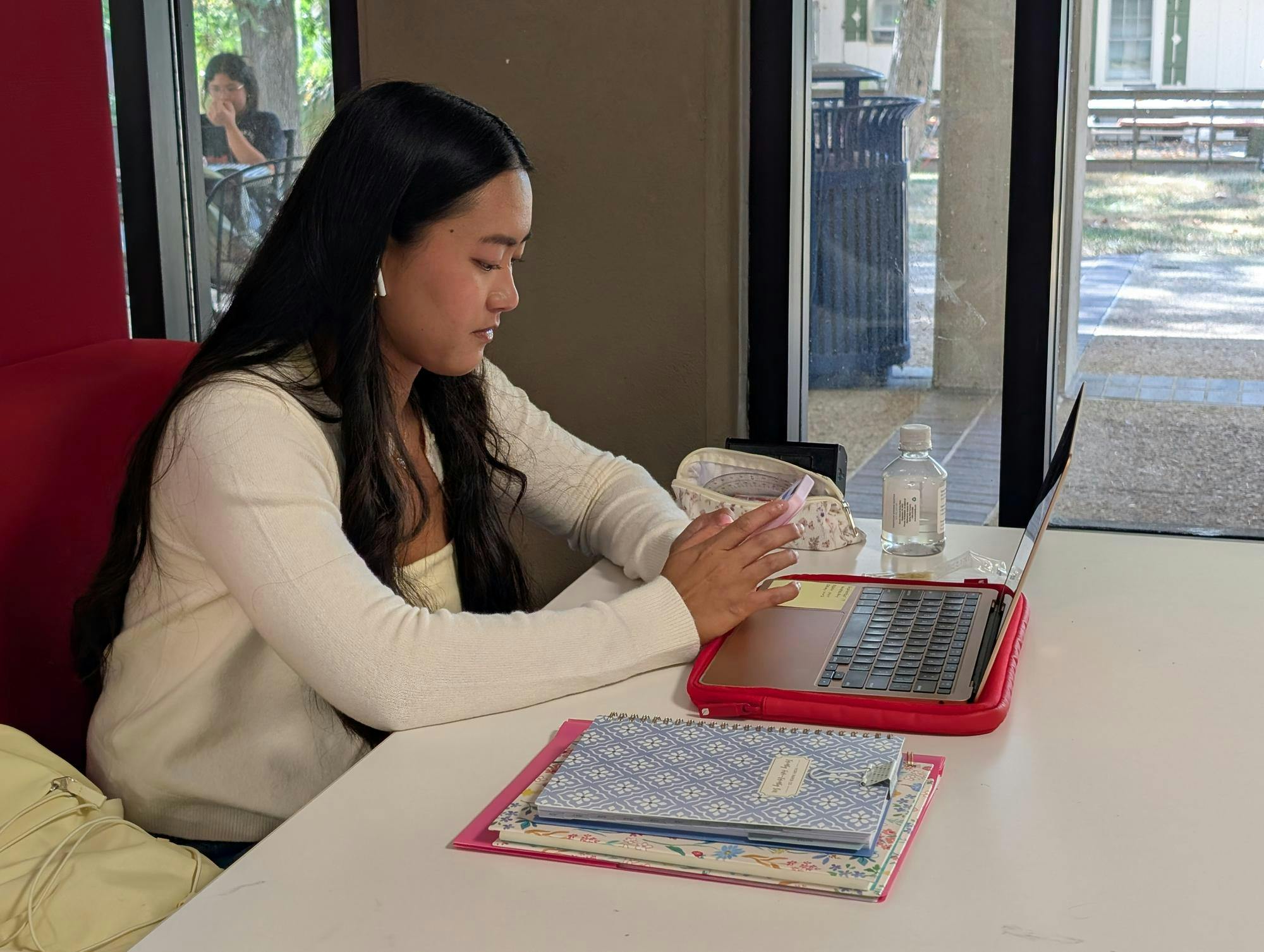 A student is sitting at a booth in the library holding her phone. There is a laptop on the table in front of her and notebooks next to her.