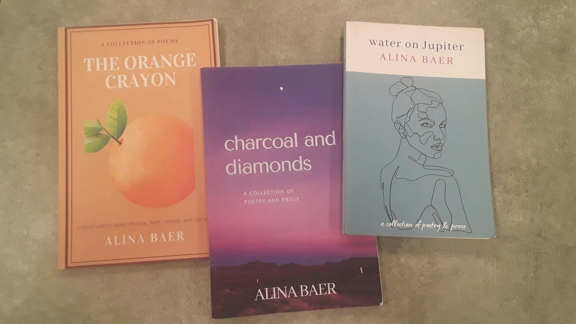 Three books by Alina Baer spread out on a table. On the left, "The Orange Crayon" features an orange. In the middle, "Charcoal and Diamonds" shows a landscape with a purple and pink twilight sky. On the right, "Water on Jupter" depicts the outline of a young woman looking over her shoulder.