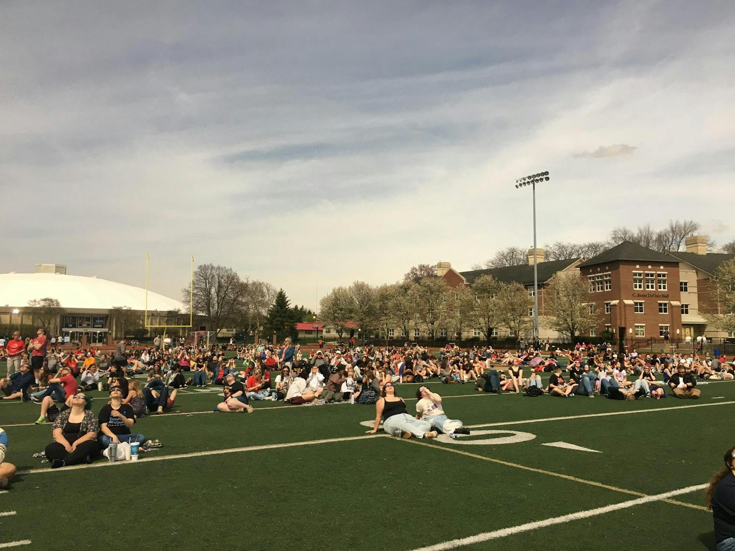 A football field filled with people sitting and laying on the grass turf, looking up at the sky with solar eclipse glasses on.