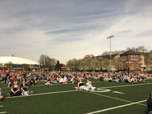 A football field filled with people sitting and laying on the grass turf, looking up at the sky with solar eclipse glasses on.