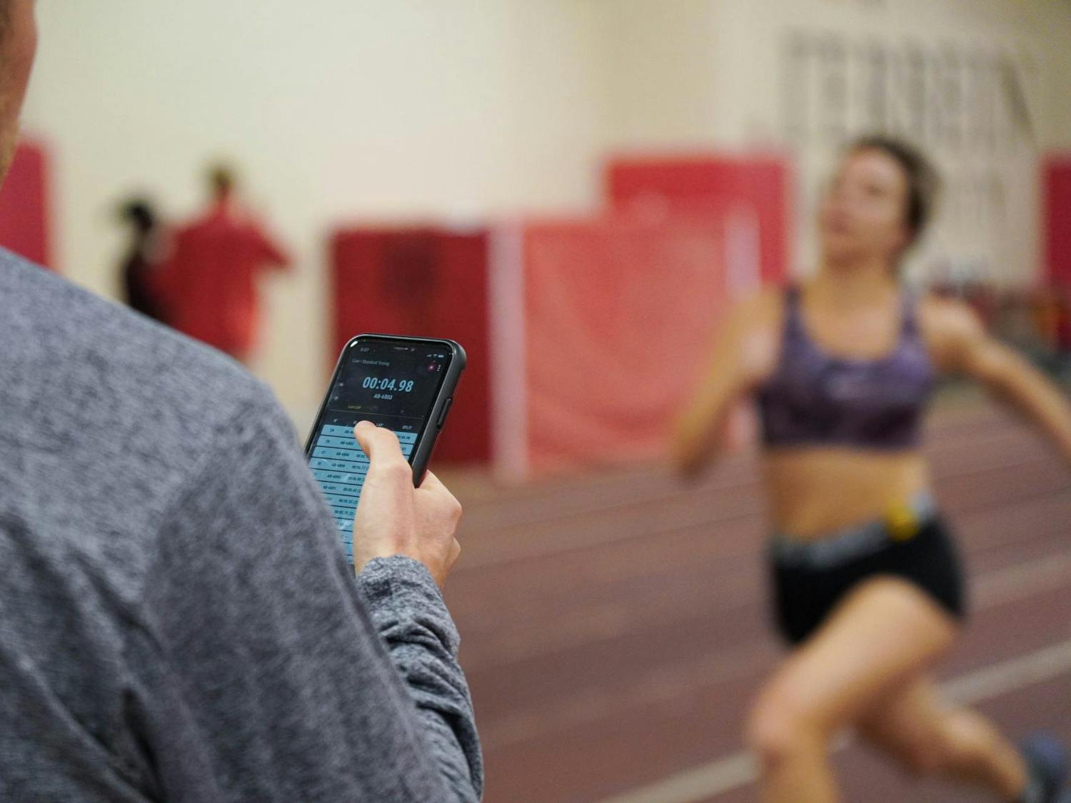 Back side of coach is positioned to the left of the frame. He holds his phone to check the student athlete's sprint time. The time reads 00.04.98. The student is blurred in the background running towards the finish line. 